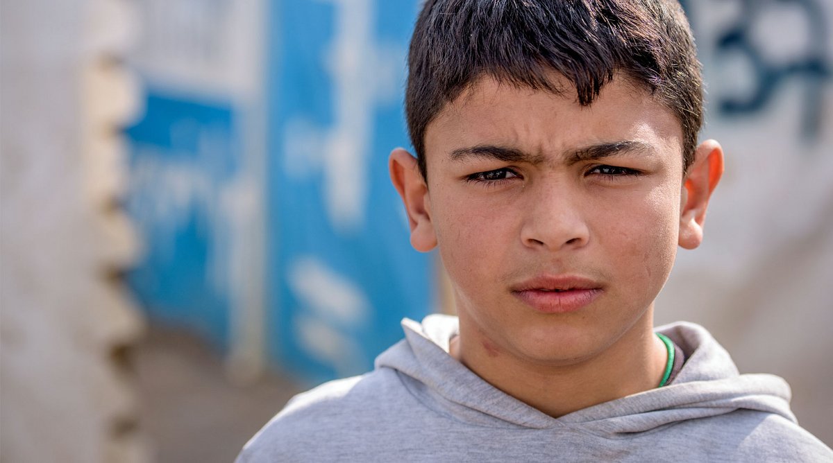 Libanon: Ein Junge in grauem Pullover blickt besorgt in die Kamera. Hinter ihm sind verschwommen Flüchtlingszelte zu erkennen.