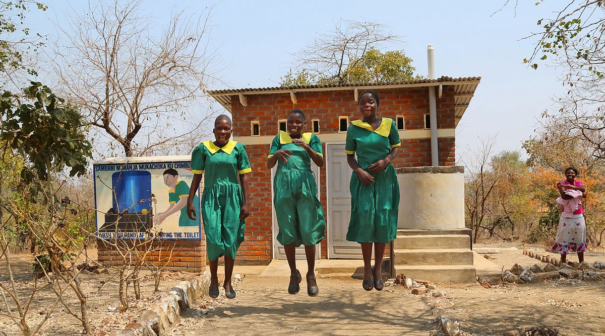 Drei Mädchen hüpfen vor Toilettenblock in Malawi.