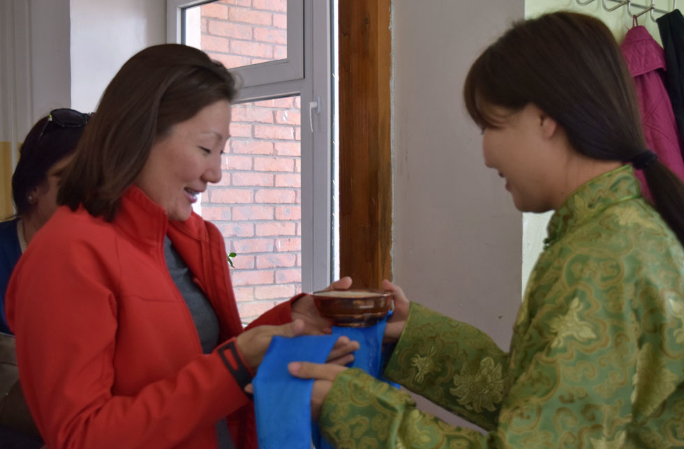 Mongolei: Eine Frau in einem traditionellen, grünen Kleid übergibt einer anderen Frau in einer roten Jacke eine Schale mit Milch und ein Tuch.
