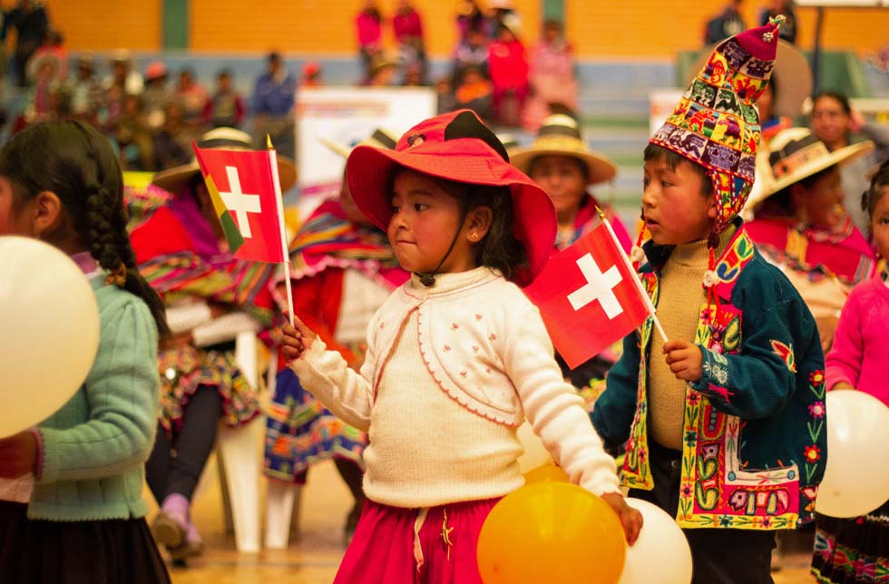 Bolivien: Kleine Mädchen und Buben in traditioneller Kleidung mit Hüten und bunten Mützen schwenken Schweizer Fähnchen.