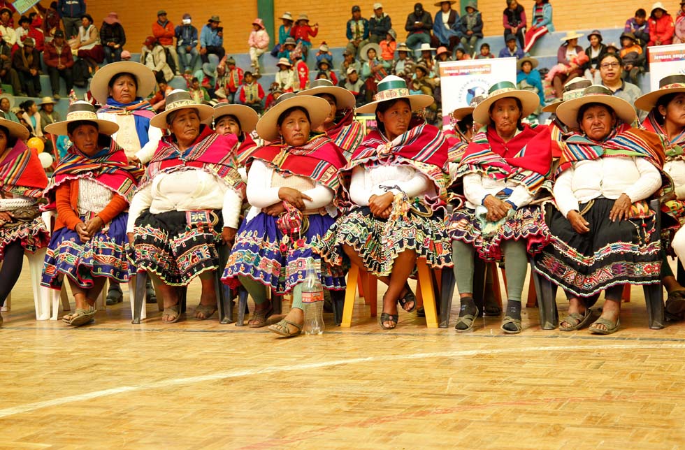 Bolivien: Eine Reihe von Frauen in traditionellen Ponchos und bunten Röcken sitzt in einem Stadion.