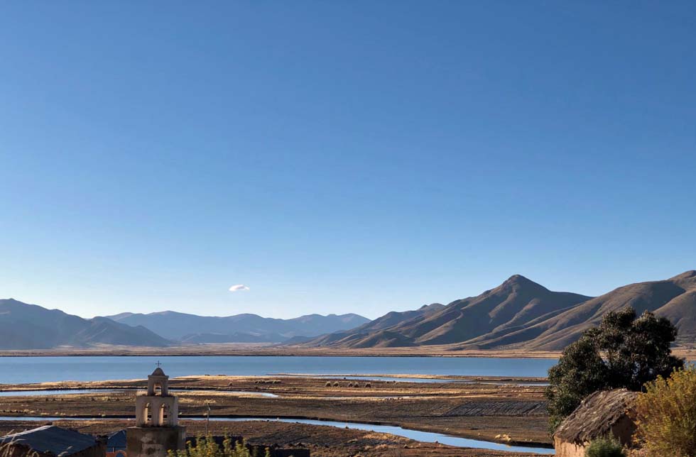 Bolivien: Weiter Ausblick auf einen Bergsee in der Mitte von Bergen, im Vordergrund ein Glockenturm.