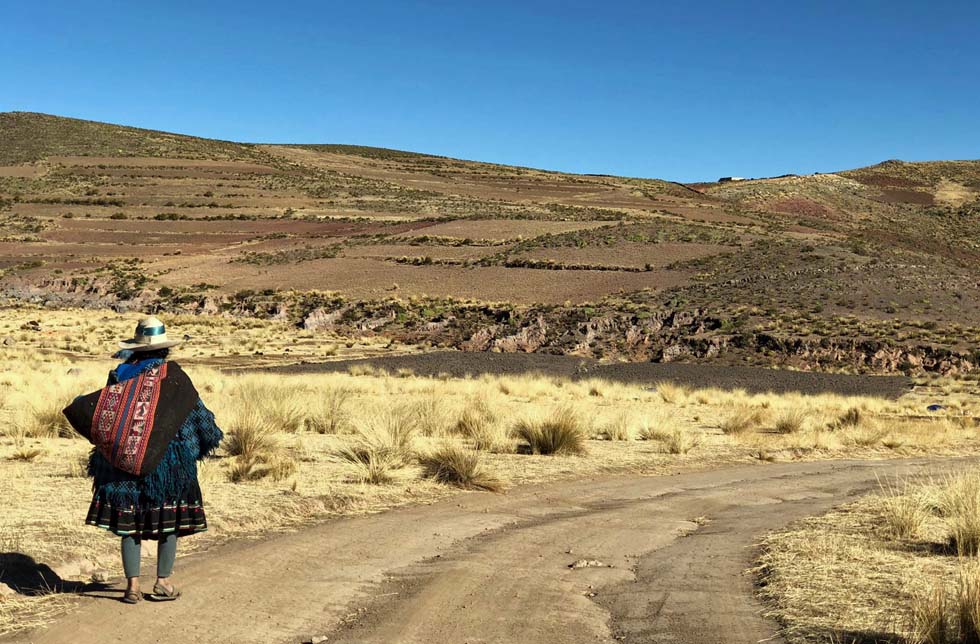 Bolivien: eine Frau in traditioneller Kleidung geht eine leere Schotterstrasse entlang. Im Hintergrund die Berge.