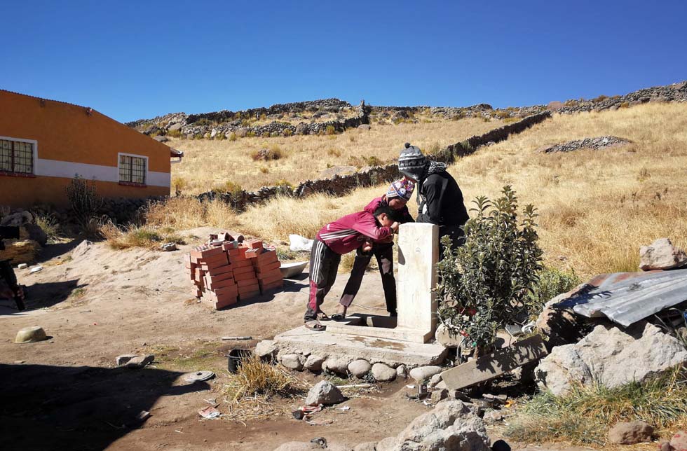 Bolivien: 3 Kinder trinken aus einem Brunnen vor einem Haus.