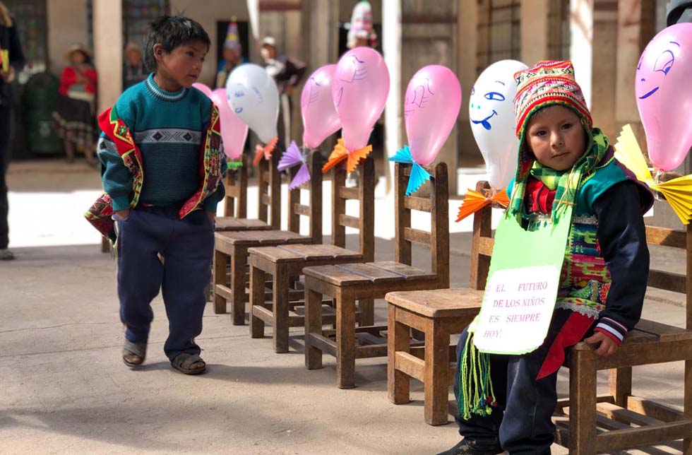 Bolivien: ein kleiner Junge sitzt am Rande einer Stuhlreihe und hat ein Schild umhängen auf dem steht: Die Zukunft der Kinder ist immer heute». Rosa Luftballons im Hintergrund