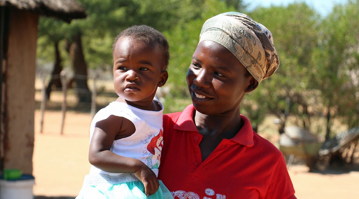 Simbabwe: Eine Mutter hält ihre kleine Tochter im Arm und blickt sie strahlend an. Hinter ihnen sind Büsche und eine Lehmhütte zu erkennen.