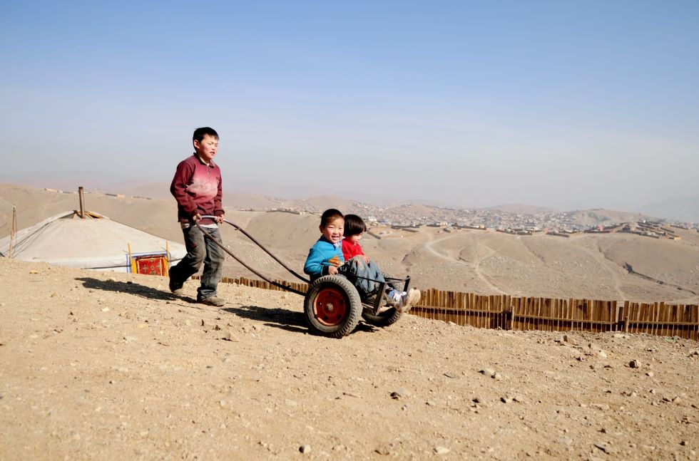 Mongolei: Ein Junge schiebt zwei kleine Junge in einem Wagen vor sich her, im Hintergrund sind karge Hügel und eine Stadt zu sehen.