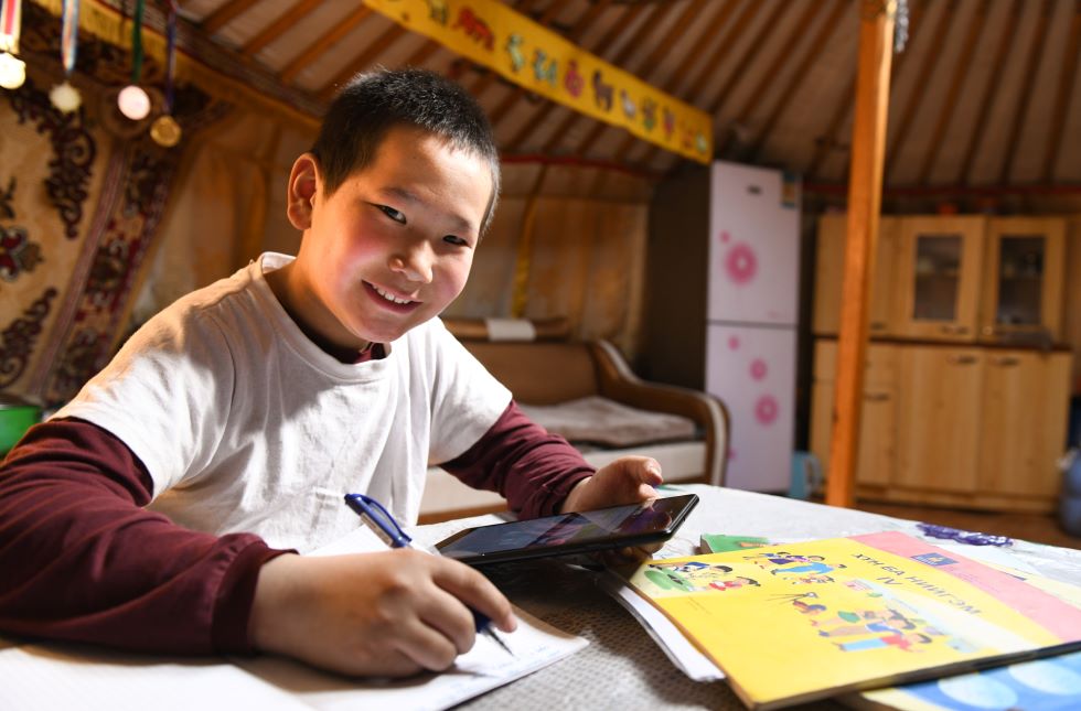 Mongolei: Ein Junge sitzt am Tisch mit Schulbüchern, er hält ein Tablet und einen stift in der Hand und lächelt in die Kamera.