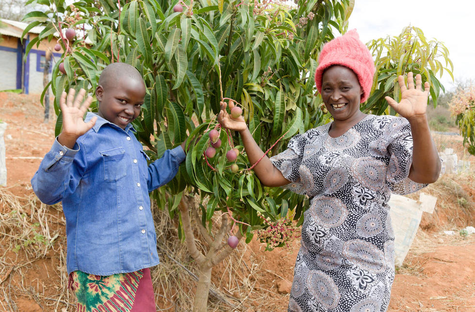 Kenia: eine Frau und ein Junge stehen vor einem Baum mit Früchten und winken lachend in die Kamera.
