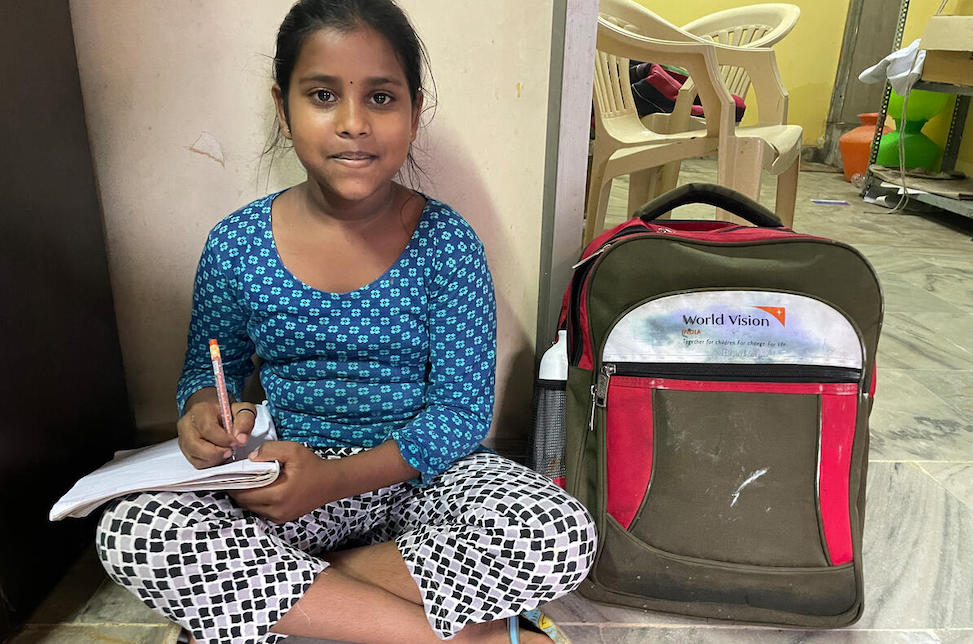  Indien: Ein junges Mädchen sitzt im Schneidersitz auf dem Boden. Auf ihrem Knie liegt ein Heft, sie hält einen Stift und neben ihr steht ein Rucksack mit dem World Vision Logo.