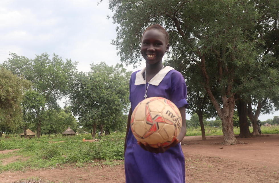 Südsudan: Ein Mädchen in Schuluniform hält mit einer Hand einen Ball in die Kamera und lächelt.