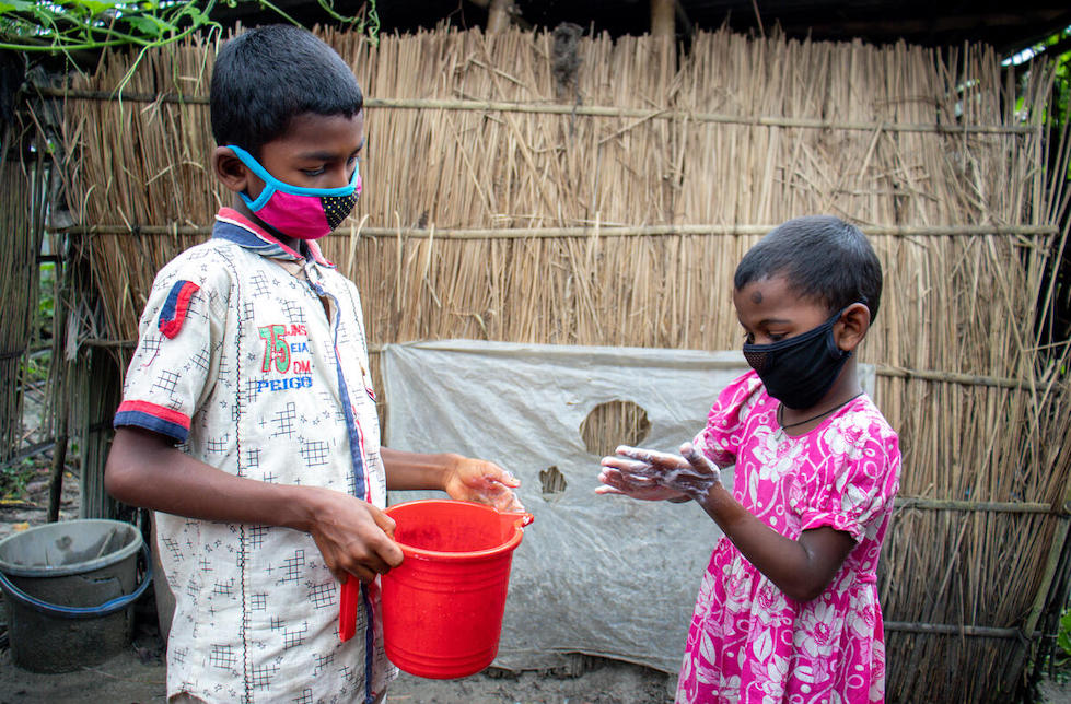Bangladesch: Ein Junge mit Maske hält einen kleinen Eimer in der Hand. Vor ihm steht ein kleines Mädchen mit Maske und seift sich gerade die Hände ein. 