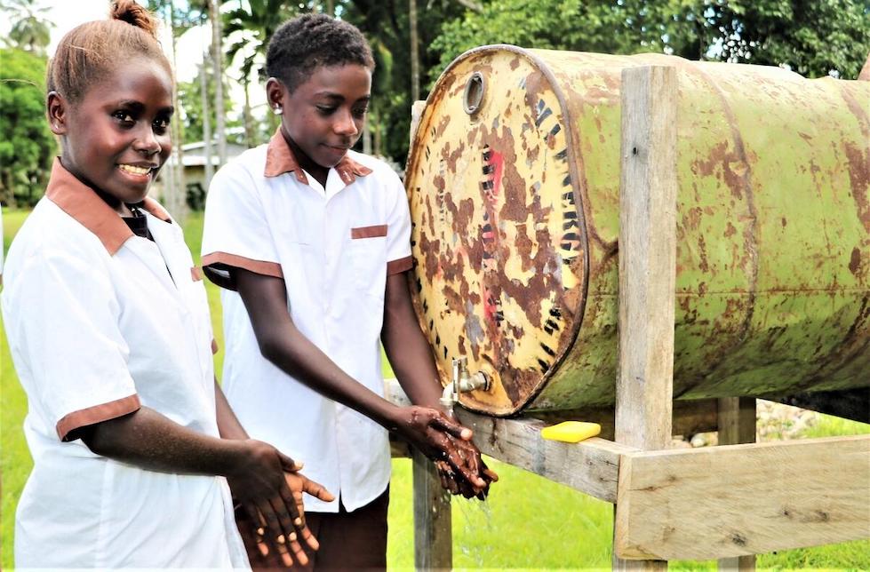 Papua Neuguinea: Zwei Mädchen in Schuluniform stehen vor einem Metallfass, in dem sich ein Wasserhahn befindet und waschen sich die Hände. Das Fass liegt auf der Seite auf einem Holzgestell. Der Wasserhahn ist in den Boden des Fasses eingebaut.