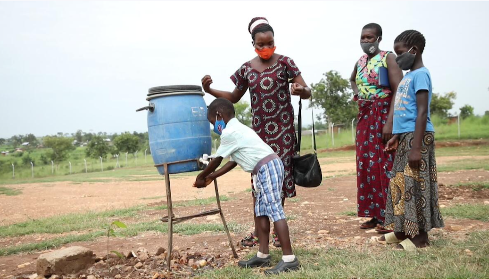 Uganda: Ein Junge wäscht sich an einer einfachen Handwaschanlage die Hände. Neben ihm steht seine Mutter und trocknet ihre Hände. Hinter ihnen steht eine Mutter und ihre Tochter und warten.