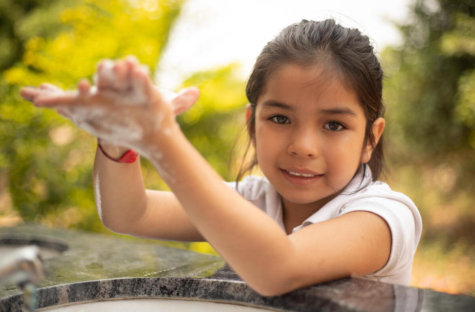 Bolivien: Ein kleines Mädchen schaut in die Kamera, während sie sich über einem Waschbecken aus Stein die Hände einseift.