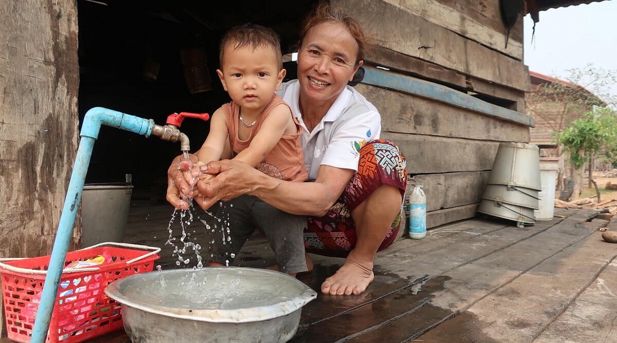 Laos: Eine Frau hockt vor einem laufenden Wasserhahn. Vor ihr steht ein kleiner Junge, dessen Hände sie mit ihren unter den Wasserhahn hält und sie wäscht. Beide blicken dabei in die Kamera.