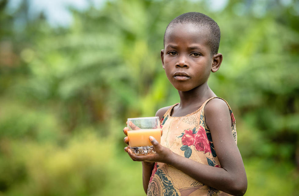 Ruanda: Ein Mädchen hält ein Glas mit einer milchig braunen Flüssigkeit in den Händen und blickt in die Kamera.