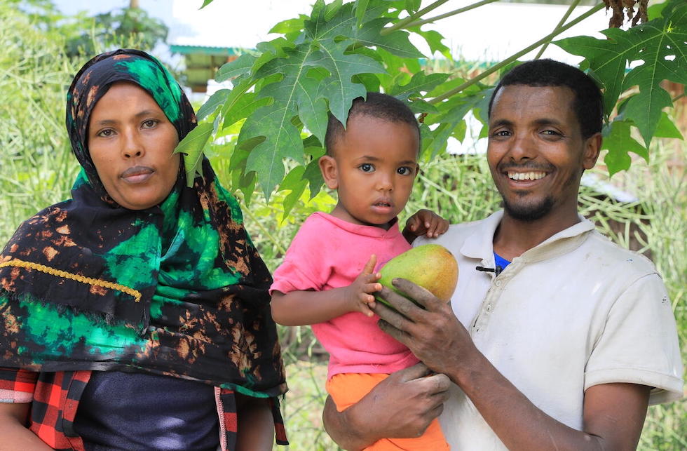 Äthiopien: Eine Frau mit Kopftuch und ein Mann mit einem kleinen Kind auf dem Arm und einer Mango in der Hand stehen unter einem Baum und blicken in die Kamera. 
