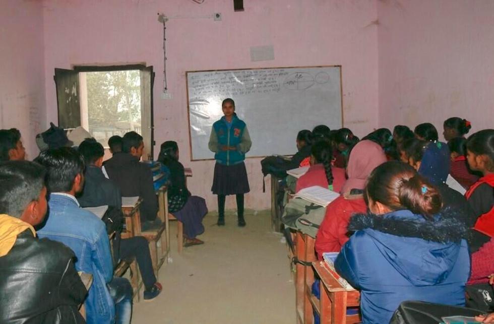 Nepal: Pinky steht in ihrem Klassenzimmer vor der Tafel und spricht zu den Kindern, die in ihren Schulbänken sitzen.