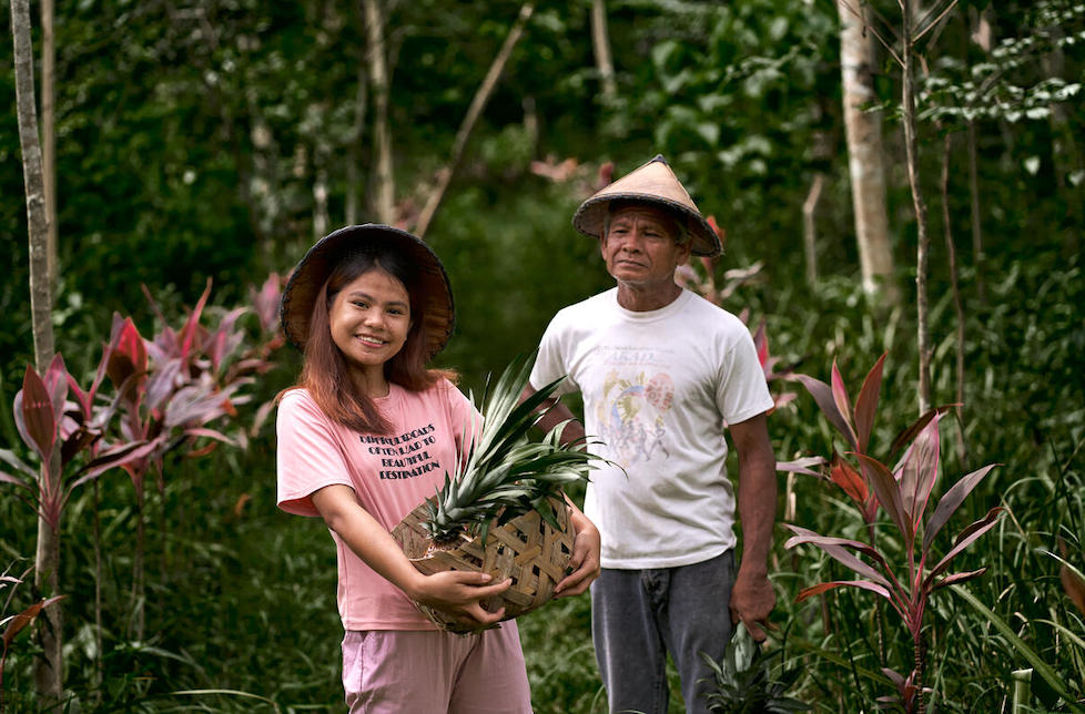 Philippinen: Ein Mädchen im pinken Shirt und mit Hut hält einen Korb mit Ananas, hinter ihr steht ein Mann und schaut sie an.