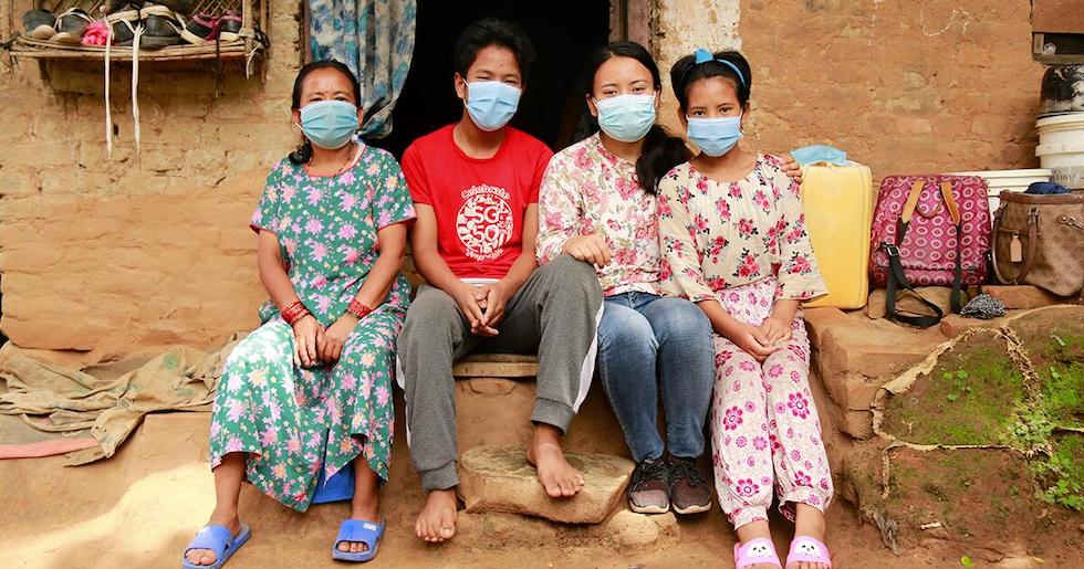 Nepal: Drei Frauen und ein Junge sitzen vor einer Hütte und schauen in die Kamera, sie tragen alle einen Mund-Nasenschutz.