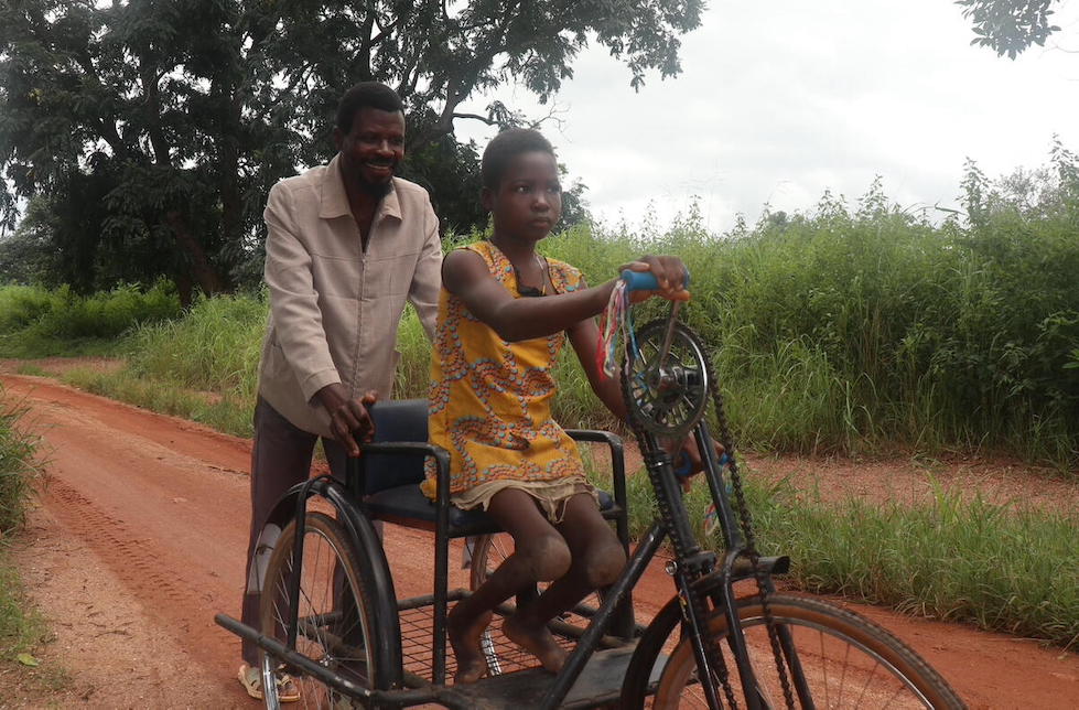 Südsudan: Ein junges Mädchen sitzt auf einem grossen Dreirad, welches sie mit ihren Händen betreibt. Hinter ihr hält ein lächelnder Mann ihre Rückenlehne.