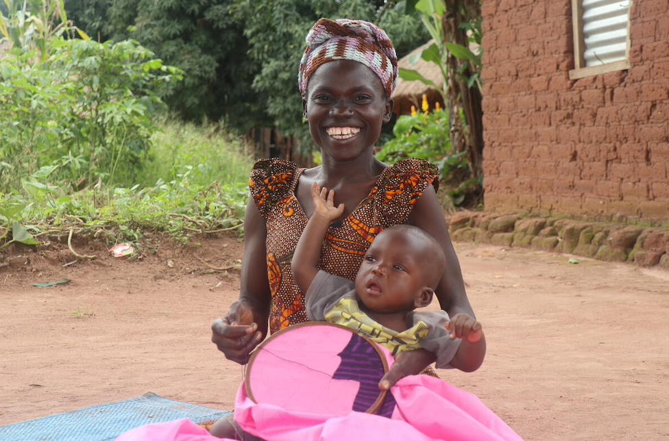 Südsudan: Eine junge Frau sitzt mit einem Baby auf dem Schoss auf dem Boden und hält einen Stickrahmen mit pinkem Stoff in den Händen.