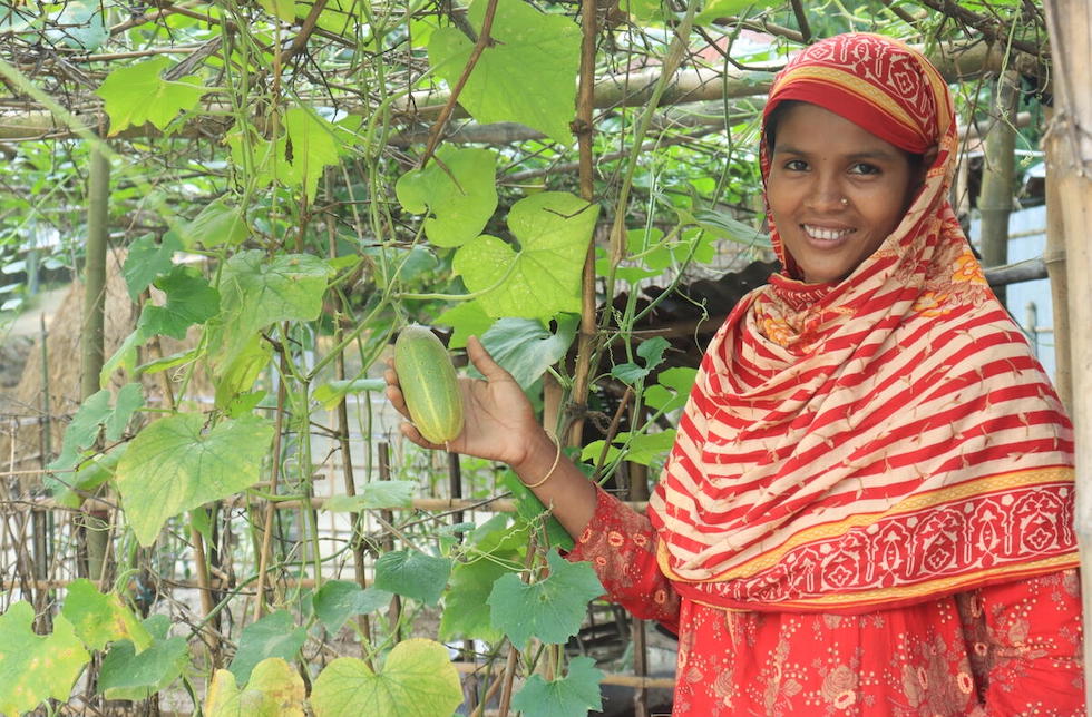 Bangladesch: Eine Frau im roten Sari schaut lächelnd in die Kamera und hält dabei Gemüse in der Hand, das neben ihr wächst.