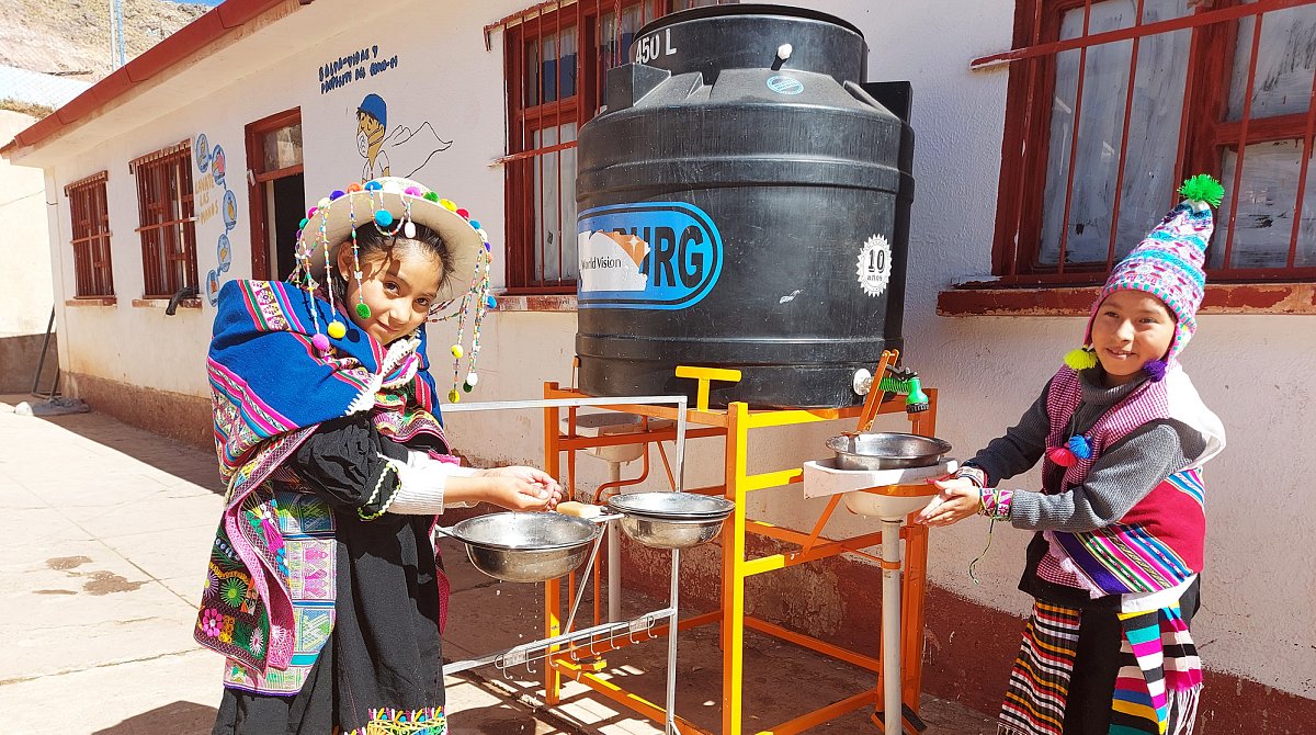 Händewaschstation: Auf einem Metallgestell im Freien steht ein grosser World Vision-Wasserkanister mit zwei Wasserhähnen, kleinen Waschbecken  und Seifenschalen. Zwei bolivianische Kinder waschen sich die Hände.
