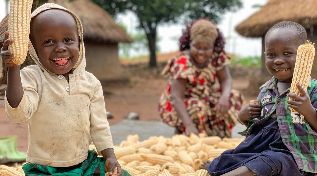 Uganda: Zwei Jungen spielen im Mais. Eine Frau arbeitet im Hintergrund