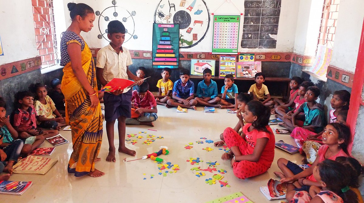 Indien: Kinder besuchen den ausserschulischen Förderunterricht. Sie sitzen mit ihren Heften am Boden. Eine Lehrerin erklärt einem Jungen eine Aufgabe.