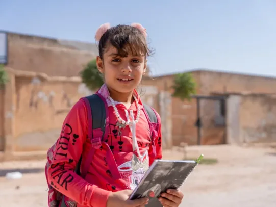 Amal lächelt im ländlichen Syrien in die Kamera, mit einem Schulheft in der Hand.