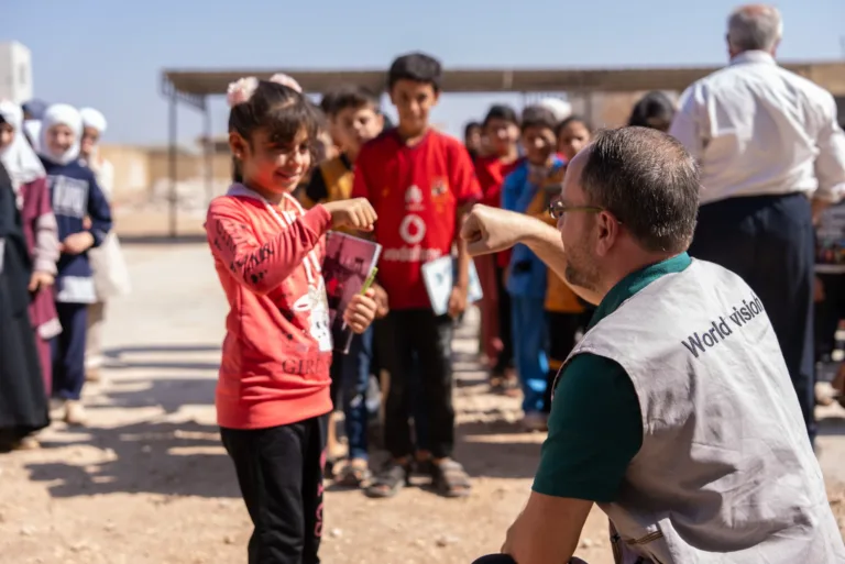 Amal begrüsst einen World Vision-Mitarbeiter im ländlichen Syrien vor dem wiederhergestellten Schulhaus. Hinter ihr stehen viele andere Kinder.